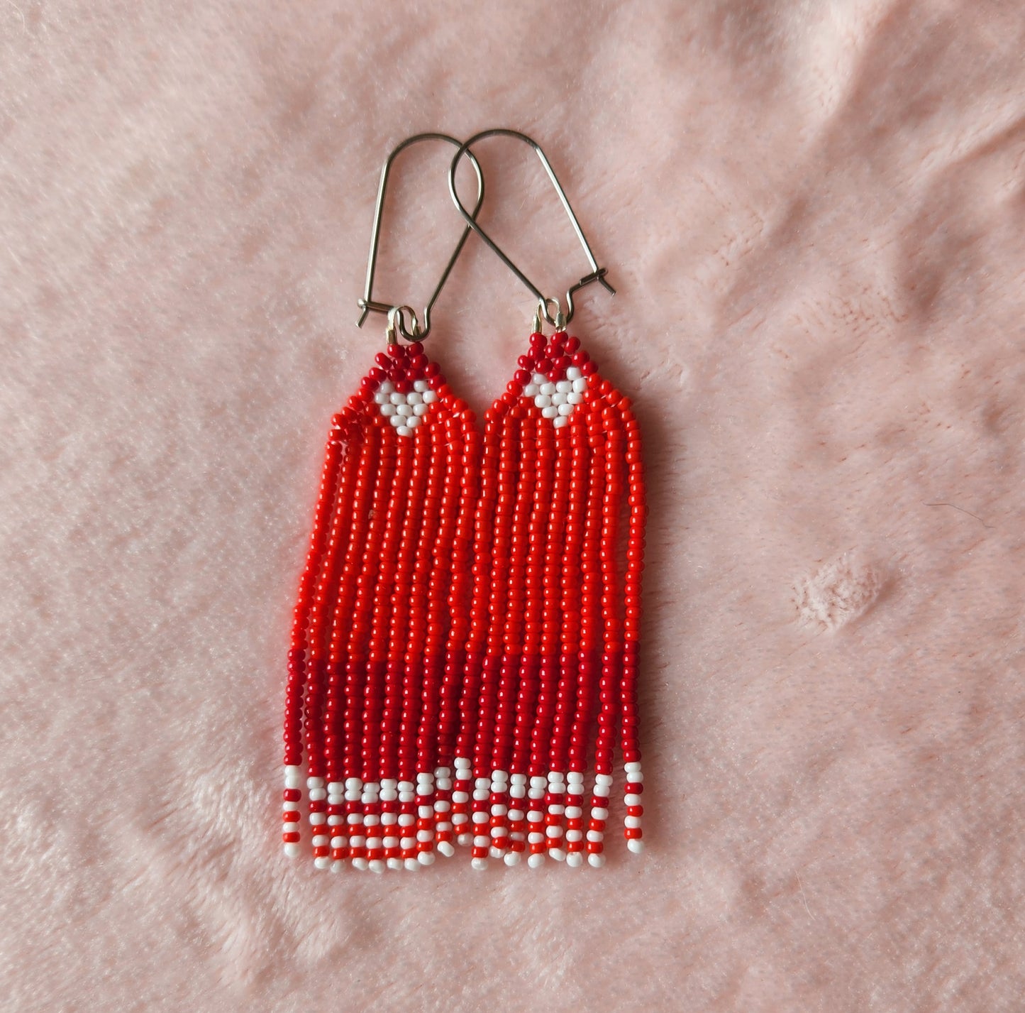 A pair of beaded fringe earrings made with varying shades of red beads, featuring a small white heart at the top and white accents at the bottom. Sat on a furry pink background.