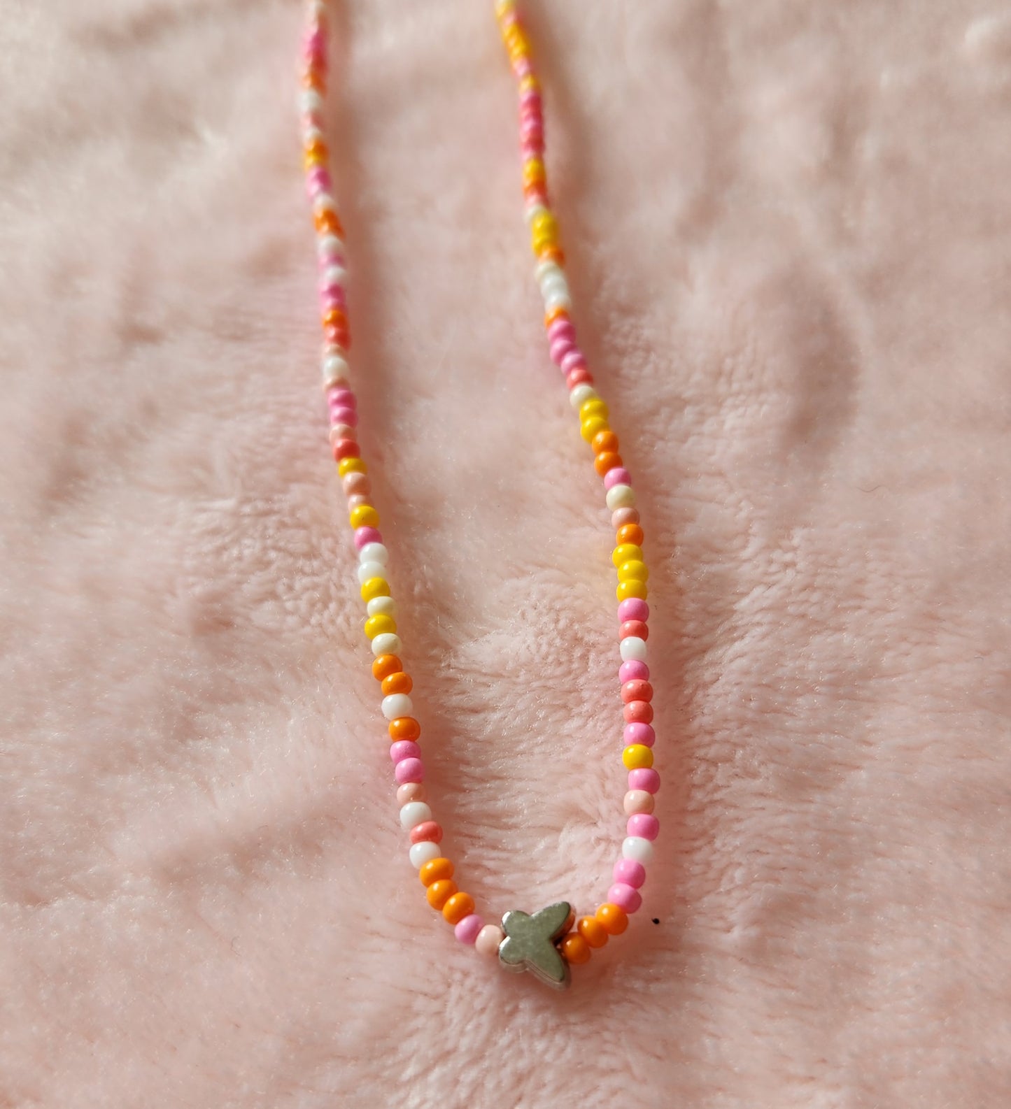 Close up of a single strand beaded anklet in orange and pink colours, with a small silver butterfly charm on a pink, furry background.