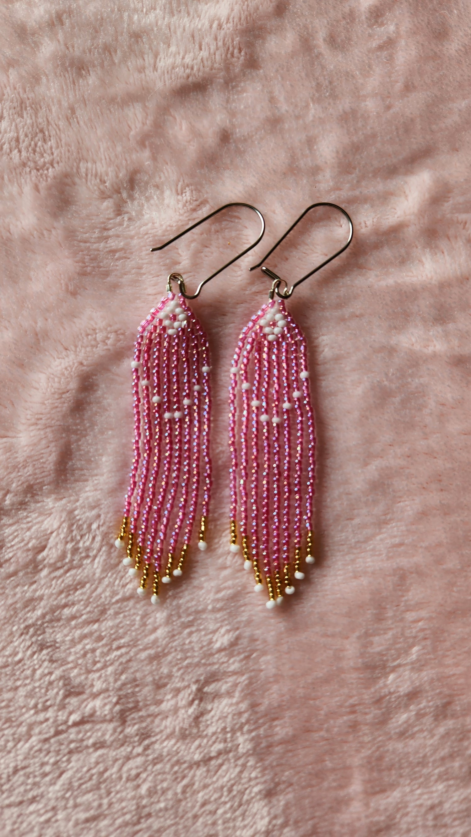 A pair of beaded fringe earrings with light pink translucent beads, with a white diamond at the top and white and gold accents on the bottom. Sat on a fuzzy pink background.