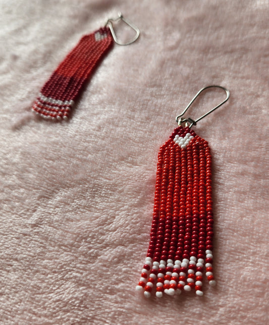 A pair of beaded fringe earrings made with varying shades of red beads, featuring a small white heart at the top and white accents at the bottom. Sat on a furry pink background. One earring is closer than the other.