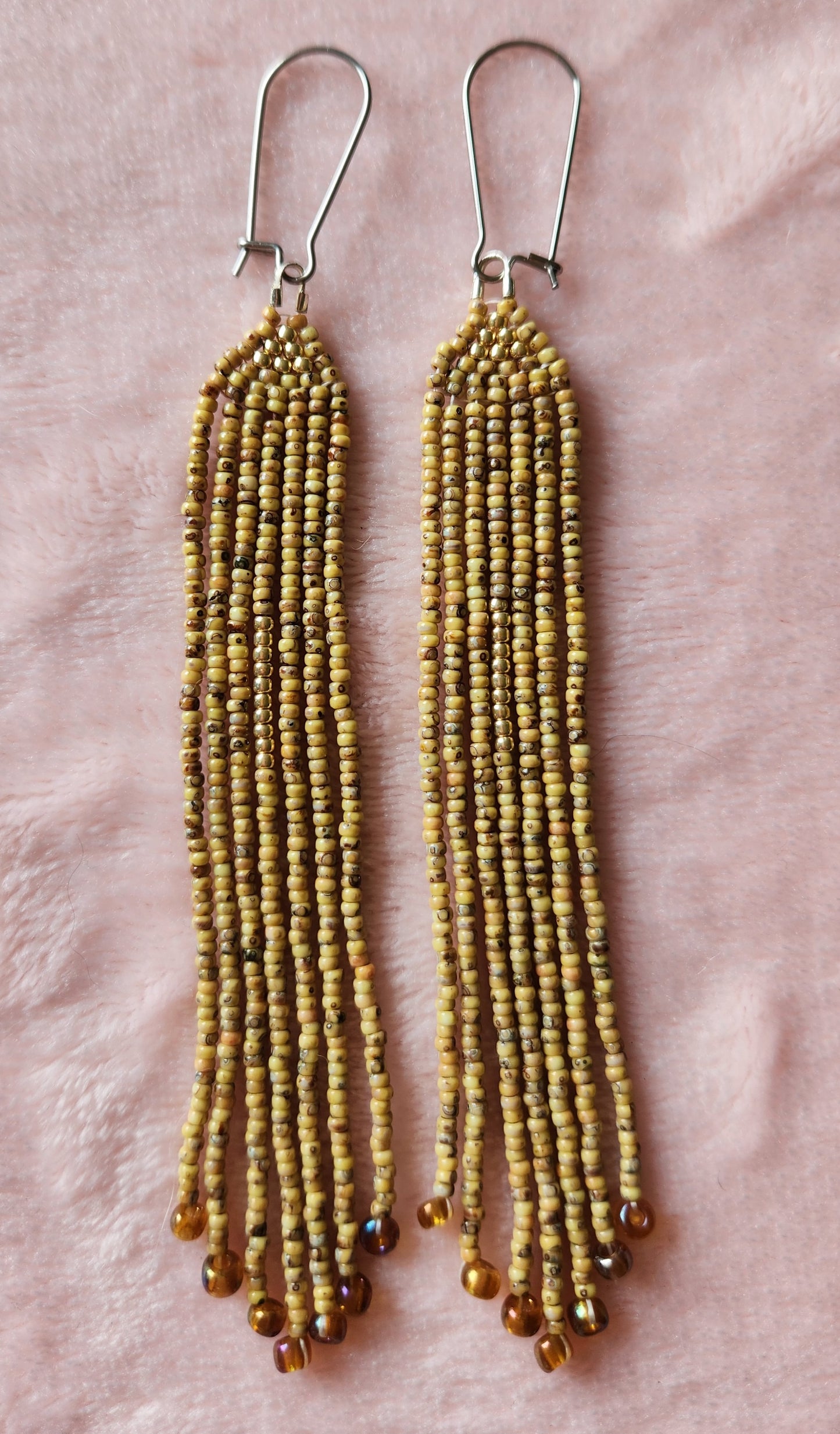 A pair of beaded fringe earrings made with brown/green coloured beads, featuring a small gold triangle at the top and copper accents at the bottom. Sat on a furry pink background. 