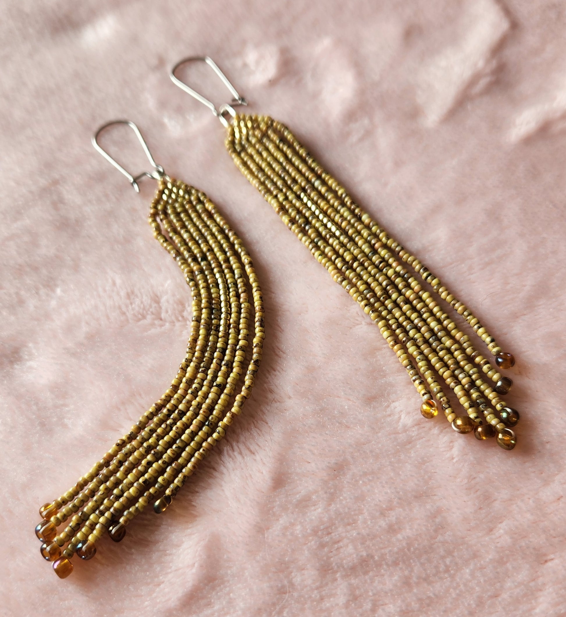 A pair of beaded fringe earrings made with brown/green coloured beads, featuring a small gold triangle at the top and copper accents at the bottom. Sat on a furry pink background. 