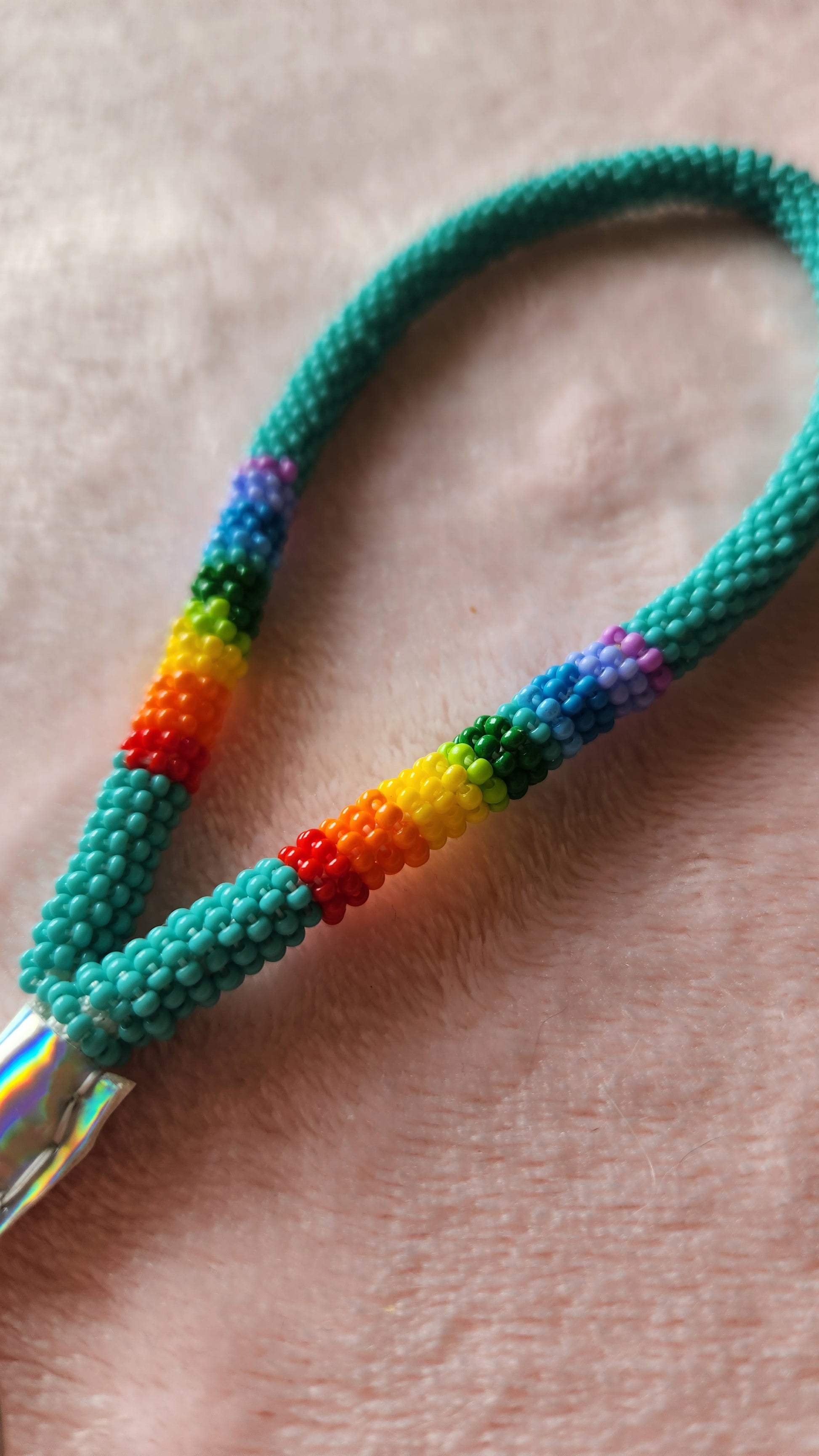 Close up of a beaded wristlet length lanyard that is mostly turquoise, with a rainbow accent and a silver faux leather wrap at the bottom. On a furry, pink background.