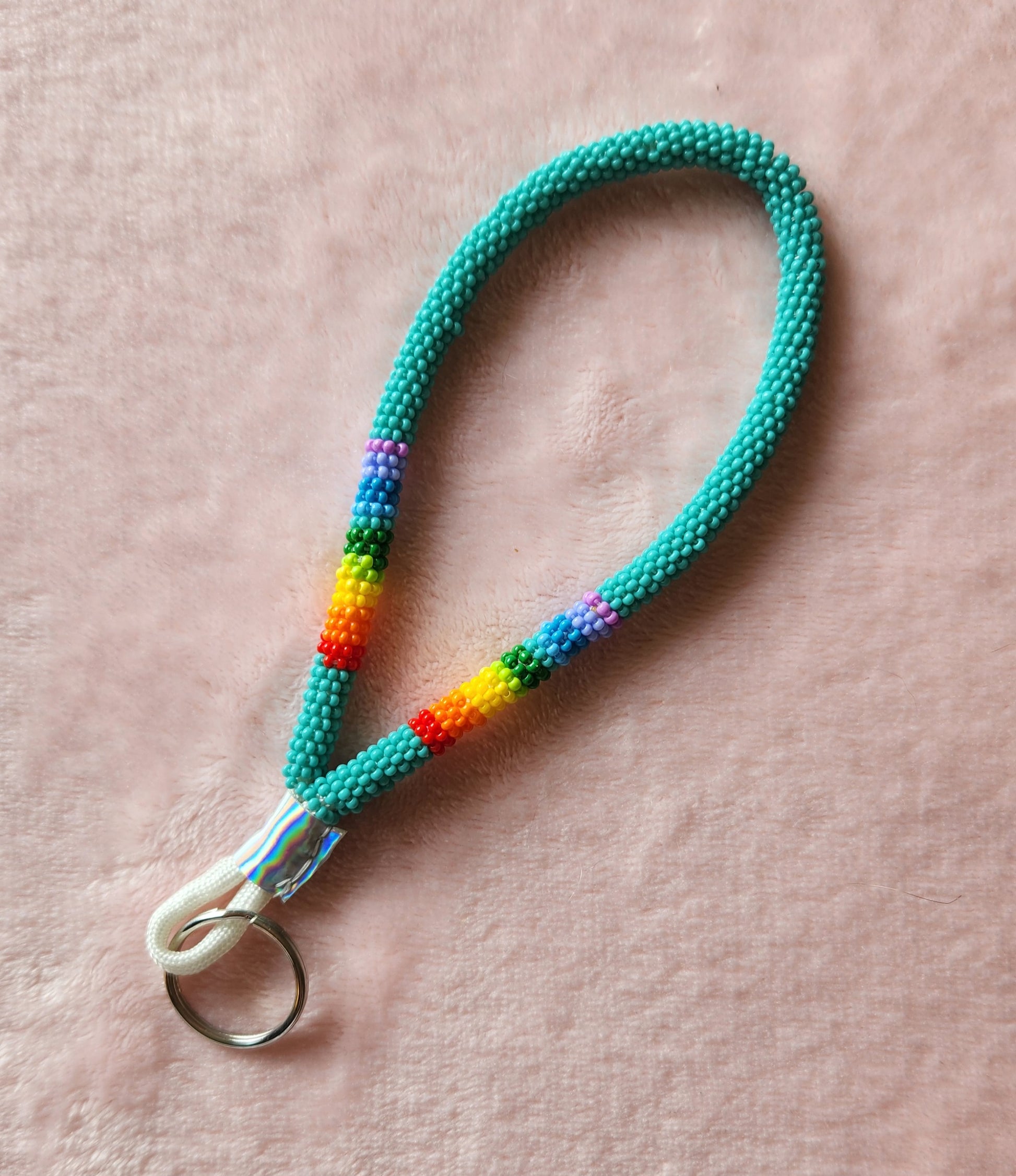 A beaded wristlet length lanyard that is mostly turquoise, with a rainbow accent and a silver faux leather wrap and keyring at the bottom. On a furry, pink background.