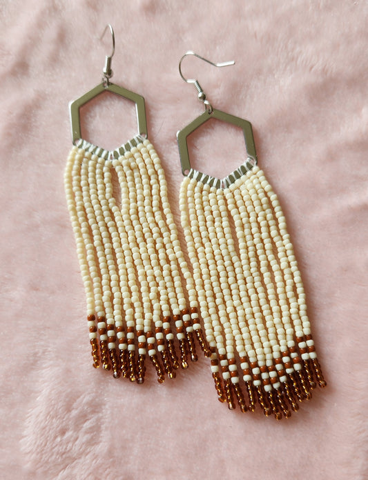 A pair of fringe earrings with cream coloured beads, and brown coloured stripes at the end, hanging off a silver hexagon shaped hoop. On a furry pink background.