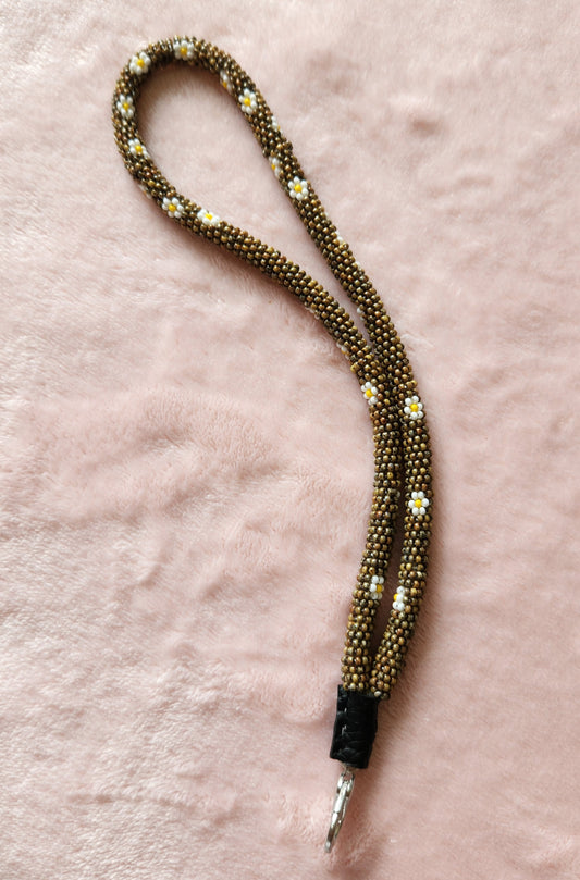 A beaded lanyard in a brown colour with white daisies on it, and a clasp at the bottom. On a pink, furry background.