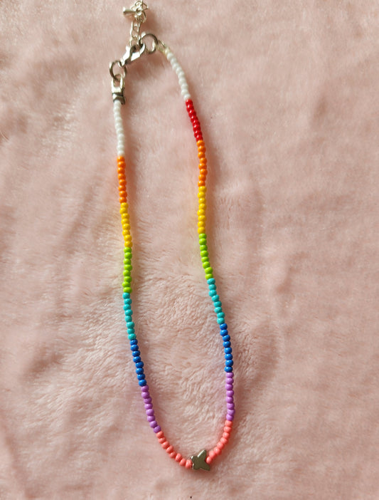 A single strand beaded anklet in a rainbow pattern with a small silver butterfly charm, on a pink, furry background.