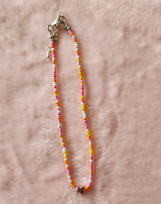 A single strand beaded anklet in orange and pink colours, with a small silver butterfly charm on a pink, furry background.