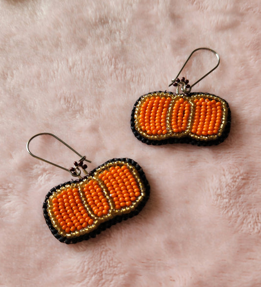 A pair of fully beaded earrings in the shape of a pumpkin, outlined in gold and filled in with orange. On a furry, pink background.