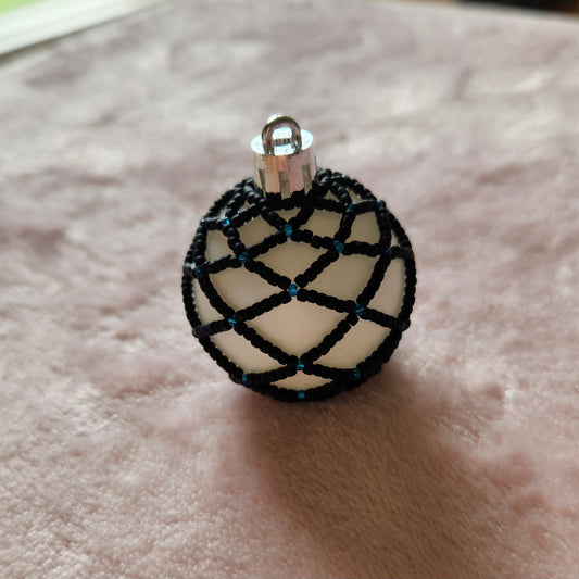 Small Christmas ornament adorned with black beads in a netted pattern, with a light blue accent on a pink, furry background.