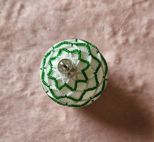 A Christmas ornament from above, adorned with beads in green and white in a striped pattern. On a furry, pink background.